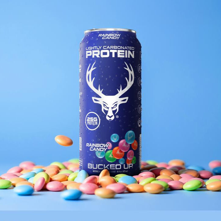 A can of Bucked Up Rainbow Candy flavored protein drink surrounded by colorful candy pieces against a blue background.